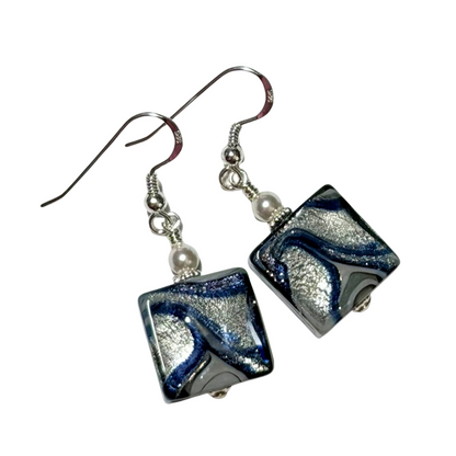 A pair of handmade square glass drop earrings featuring a marbled swirl of dark blue, silver, and black, suspended from silver fishhook wires with small white pearl beads.